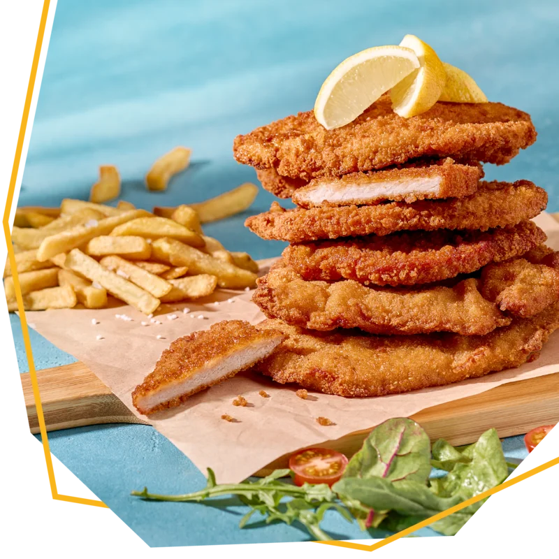 Frontansicht von gestapelten Schnitzeln auf einem Brett. Obendrauf liegt eine Zitrone und im Hintergrund Pommes.