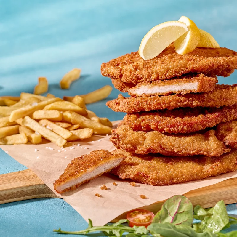 Frontansicht von gestapelten Schnitzeln auf einem Brett. Obendrauf liegt eine Zitrone und im Hintergrund Pommes.